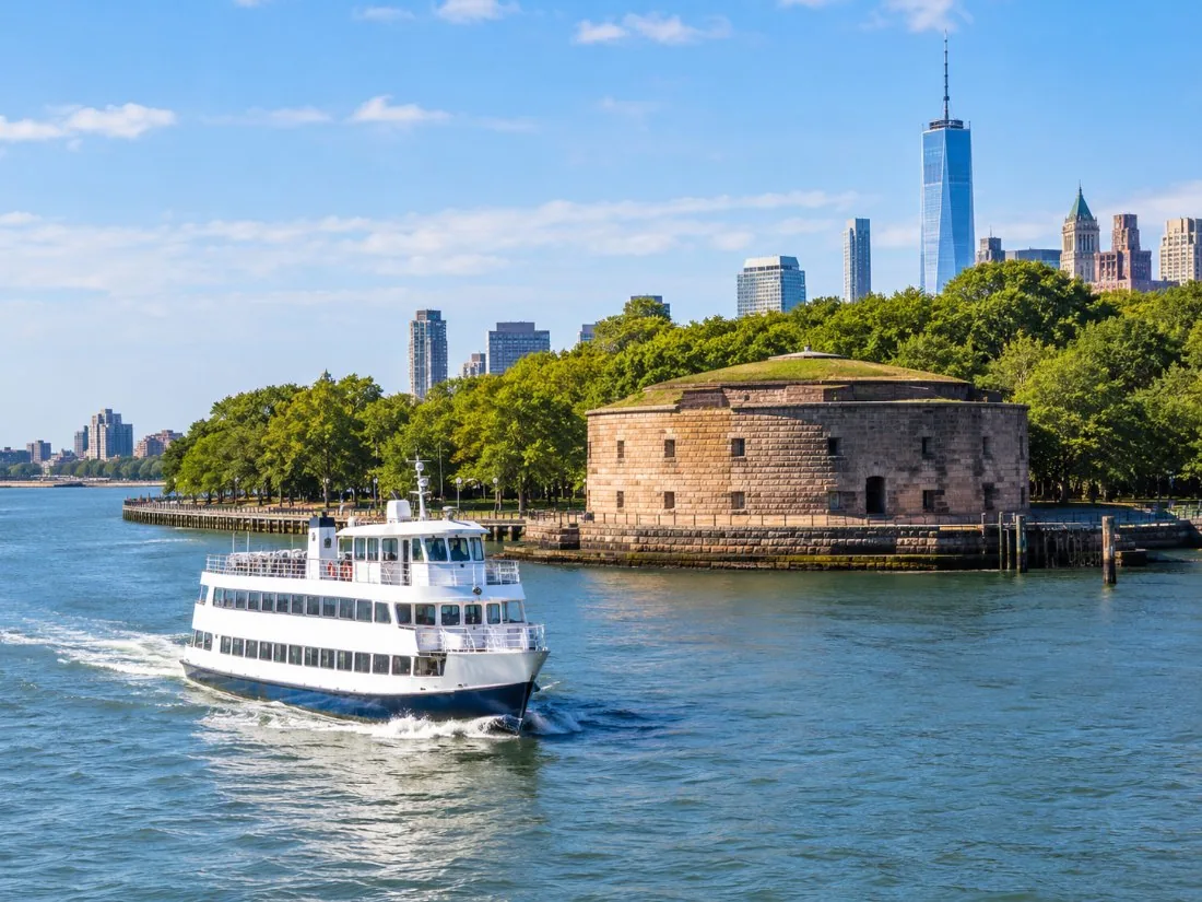 A white double-deck ferry pulling away from a stone fort at the tip of Manhattan on a sunny morning