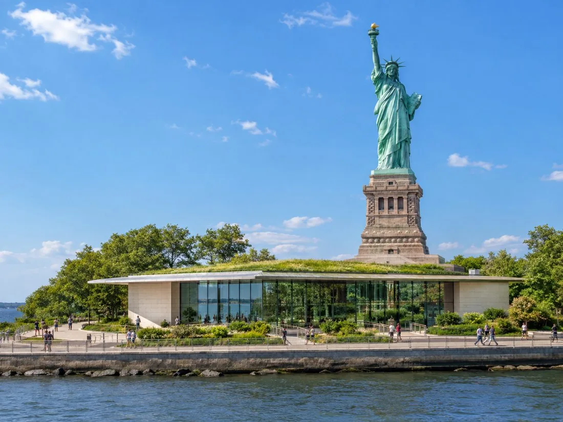 A low glass and granite museum pavilion on Liberty Island with the statue standing behind it