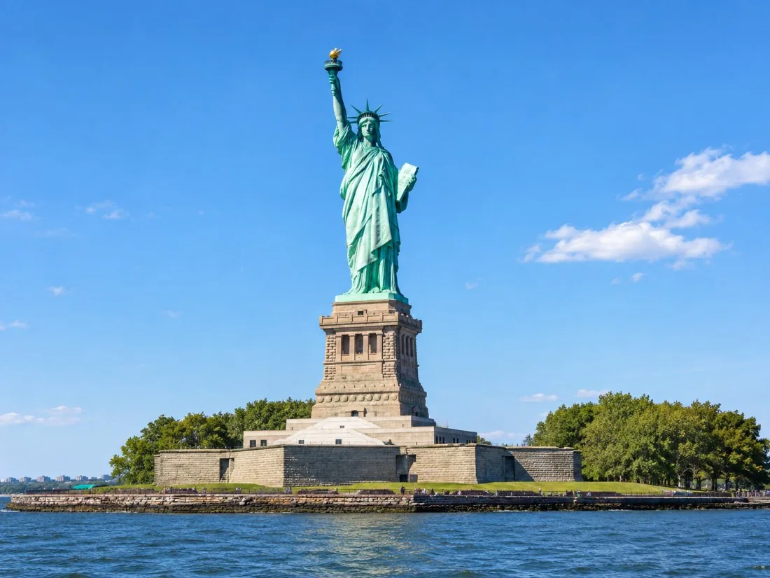 The Statue of Liberty, green copper against a clear blue sky, seen from the water with the torch raised